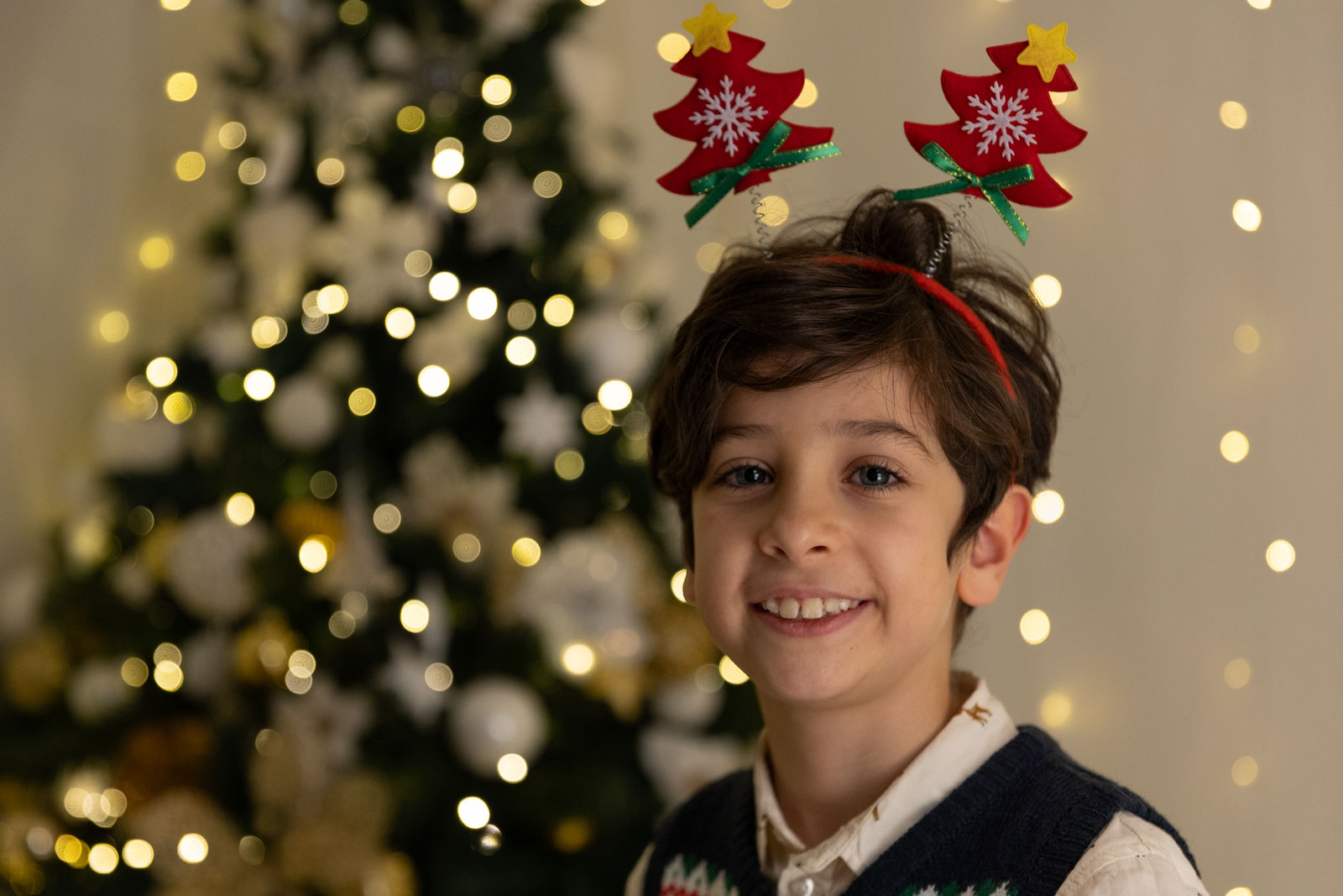 Bambino con occhi azzurri e antenne natalizie con sfondo un albero di Natale